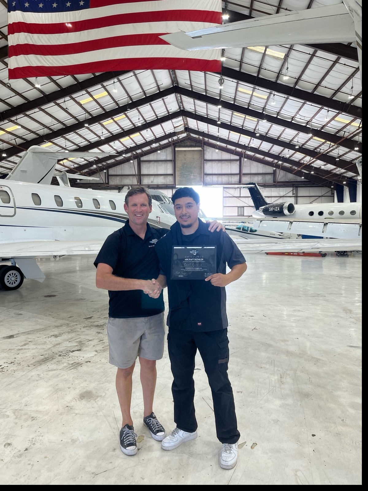Saul Garcia receiving his Shiny Jets Aircraft Detailer certification from instructor Brett Berry, in a hangar with business jets behind them.
